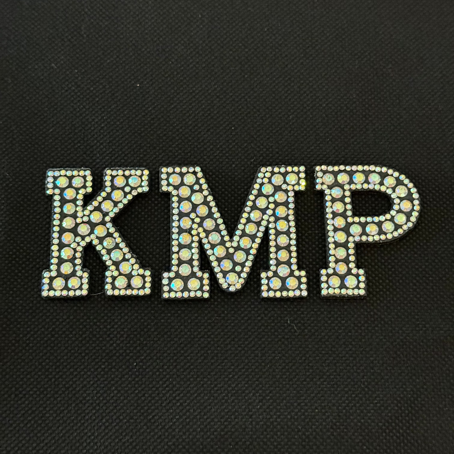 Decorative letters 'KMP' with rhinestones on a black background