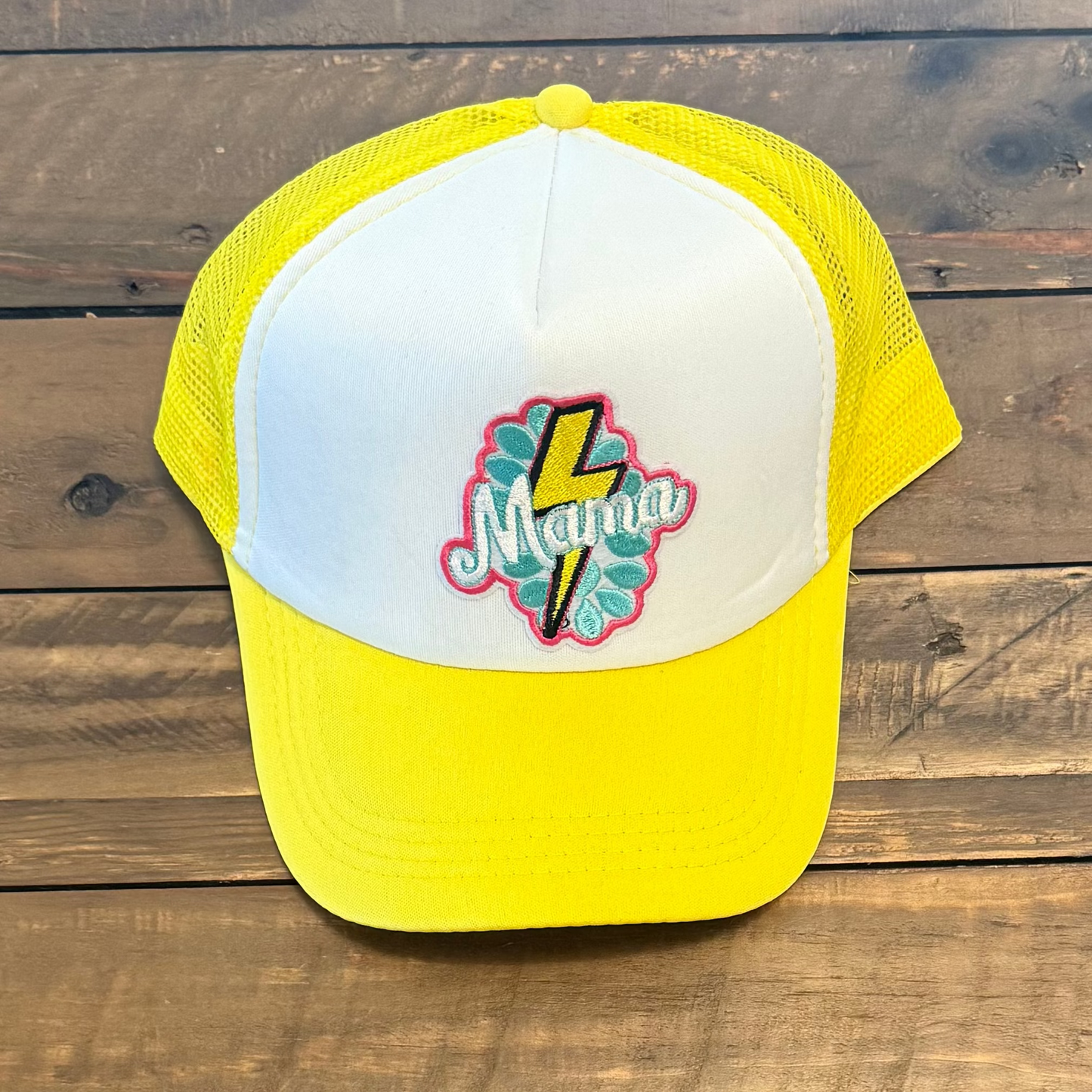 Yellow and white cap with colorful design patch that says "Mama" on a wooden surface