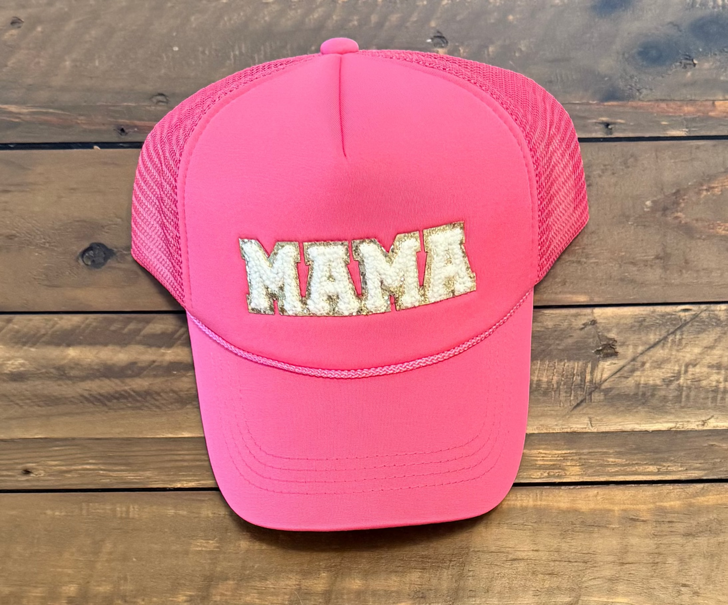Hot pink hat with pink rope detail along the brim with bold, capital, gold outlined, white, fuzzy patch lettering saying "MAMA" Pic taken on a wooden background.