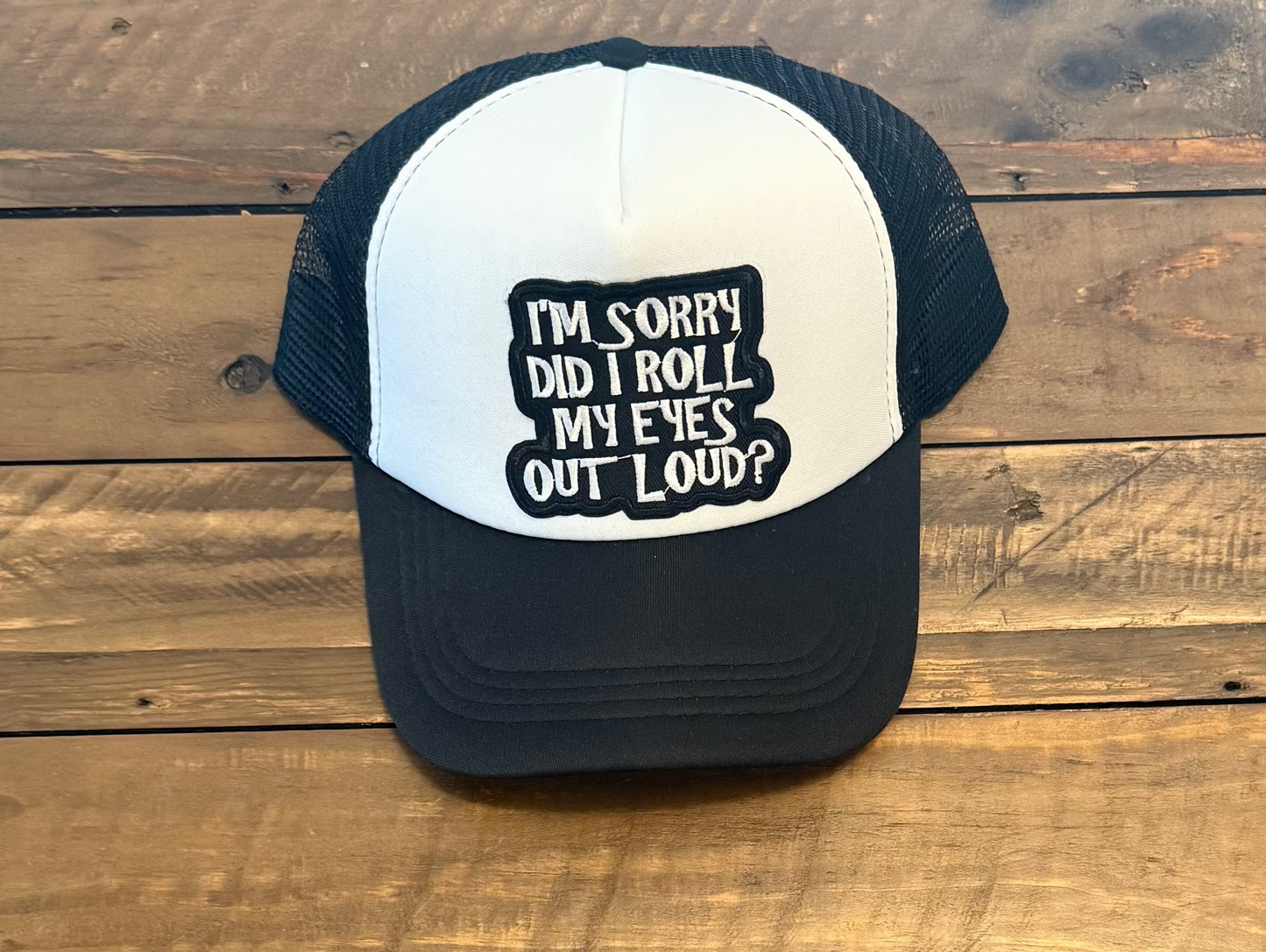 Black and white cap with a black and white text patch in the middle saying "I'm sorry did i roll my eyes out loud?" pic taken on a wooden surface.