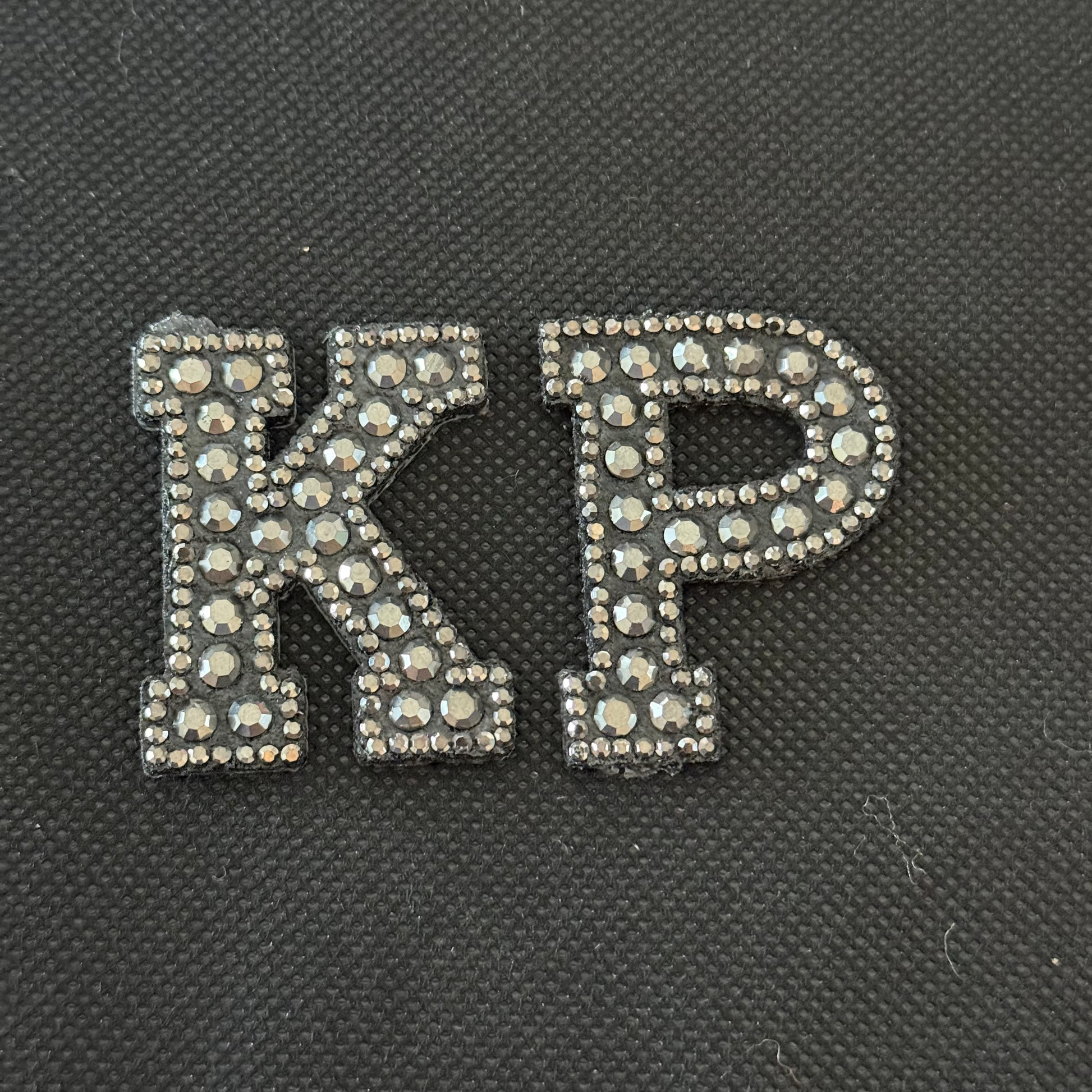 Decorative letters 'KP' with rhinestone embellishments on a black textured background