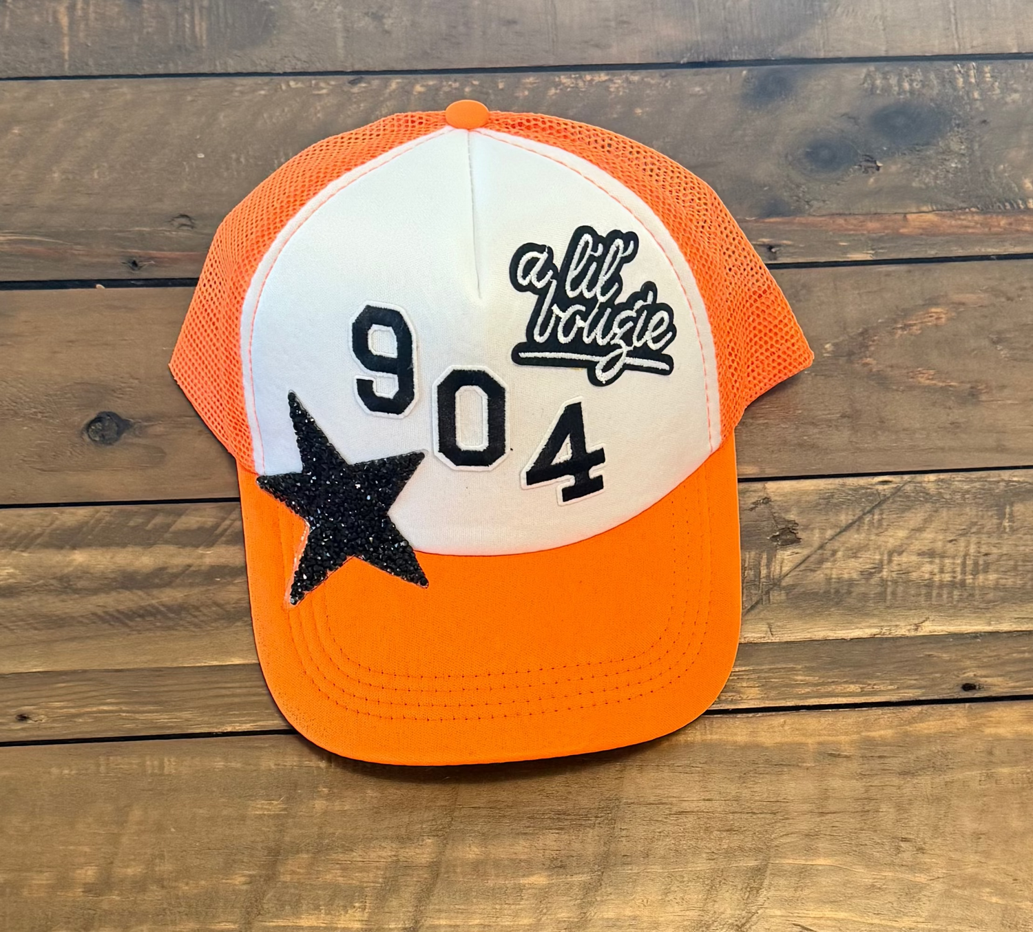 Orange and white hat with black text patches that say "904" and "a lil bougie" Also a black star patch. photo taken on a wooden backdrop