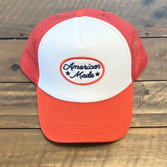 Red and white cap with a patch in the center that says "American Made" in black cursive letters with a black star on either side of the word 'Made". Photo taken on a wooden background. 