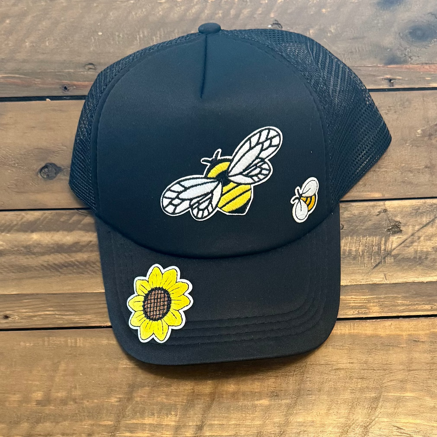 Black cap with a sunflower patch and 2 bumble bee patches on a wooden surface