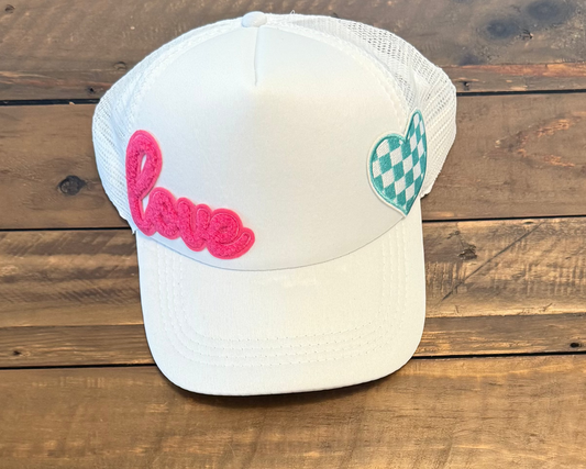 White cap with a pink cursive fuzzy "love" patch on the left and a blue and white checkered heart patch to the right of the hat. pic taken on a wooden surface.
