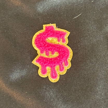Pink dollar sign patch with gold outline on a dark fabric background