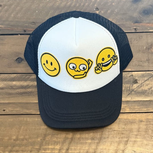 Black Trucker cap with smiley face emoji, Hands to the side emoji, and a smiling emoji with peace sign hands. Hat is placed on a wooden surface background. 