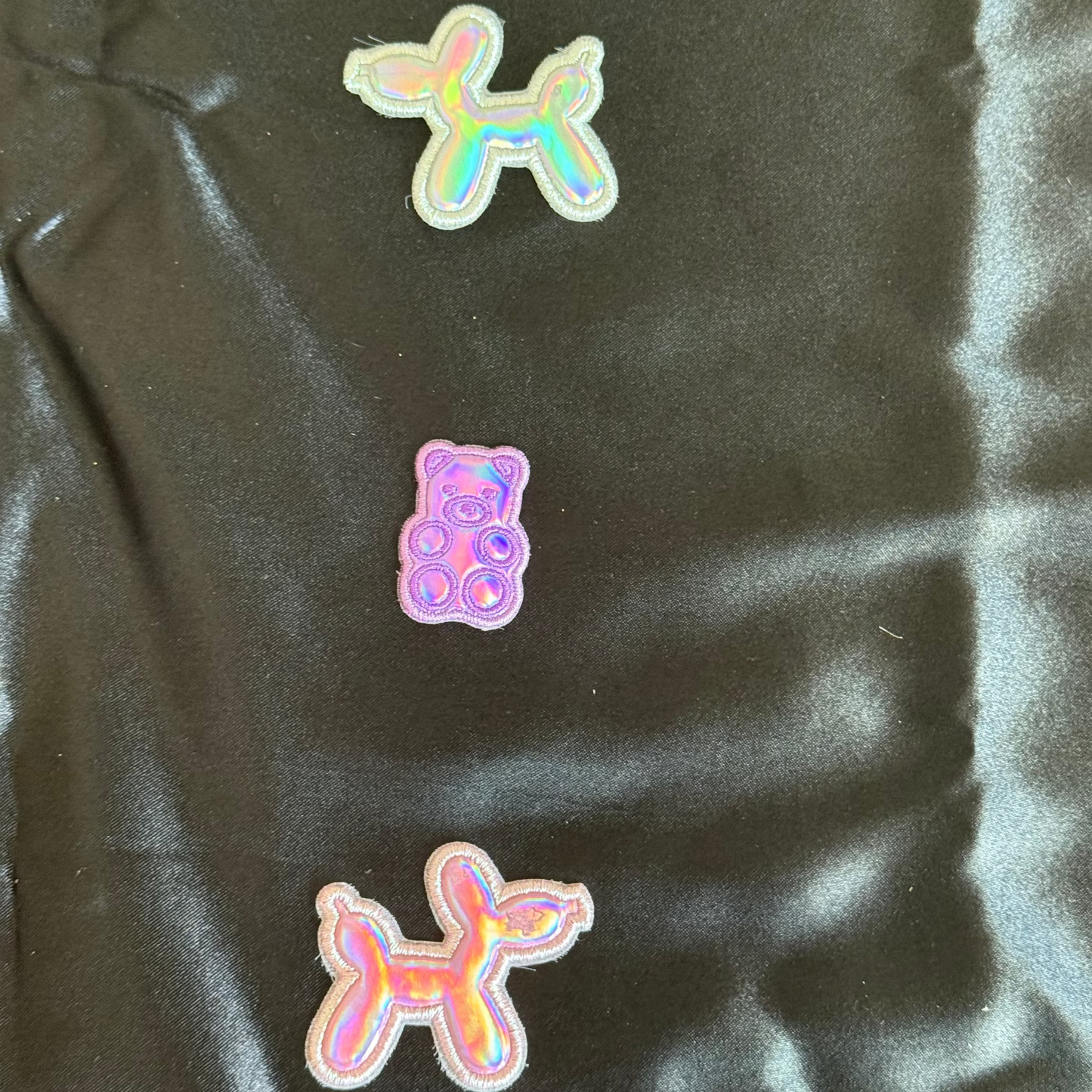 two holographic balloon dog-shaped patches and one holographic bear patch on a black satin surface