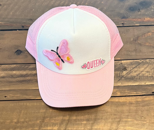 Pink and white cap with a pink butterfly patch and a patch that says "Queen" in pink lettering with pink hearts on either side of the word. Pic taken on a wooden surface