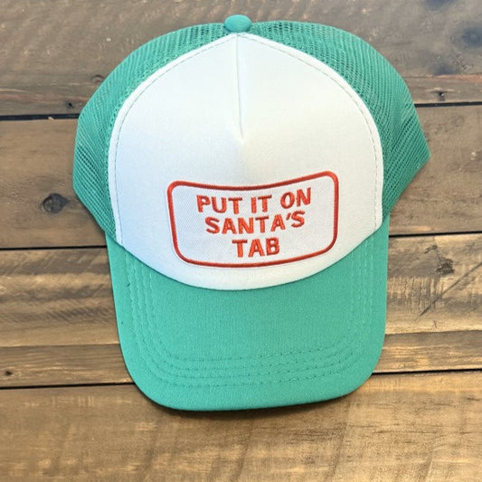 Green and white mesh trucker cap with white and red logo "Put it on Santa's Tab". Hat is placed on a wooden surface.